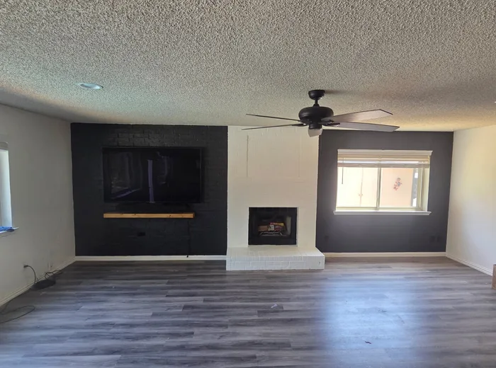 Renovated living room with a charcoal painted brick fireplace wall, mounted TV, floating wood mantel, and new grey plank flooring